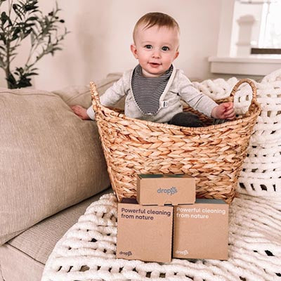 Child in a laundry basket with three boxes of Dropps
