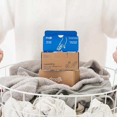 A full laundry basket being carried with two boxes of Dropps 