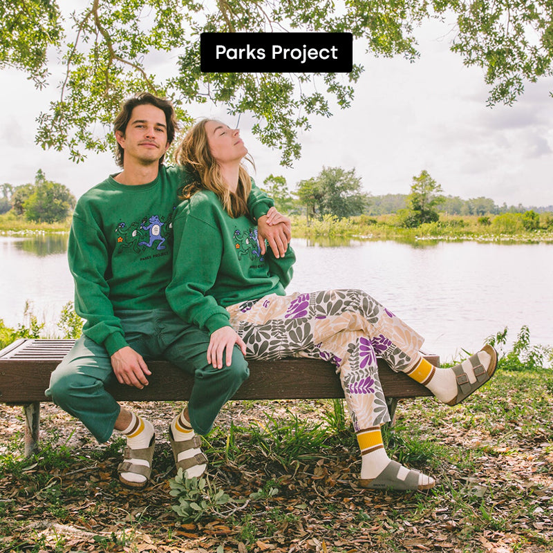A man and woman sitting on a park bench wearing Parks Project merchandise